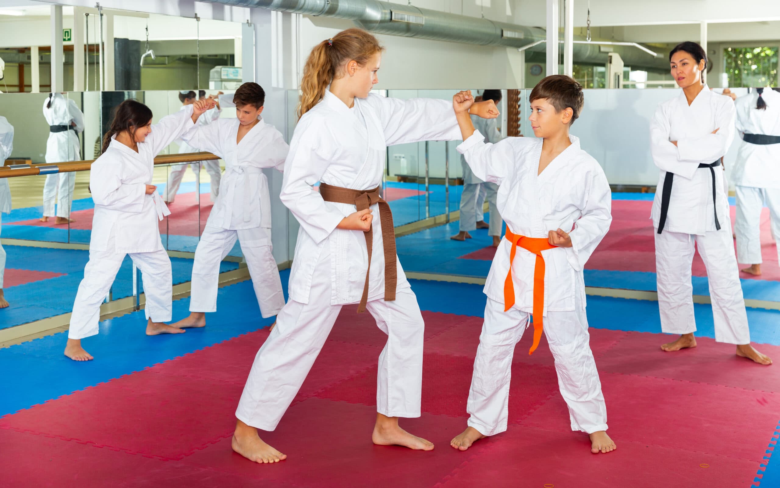 Children in karate uniforms practice blocking techniques with partners during a martial arts class while an instructor observes their form in the dojo.