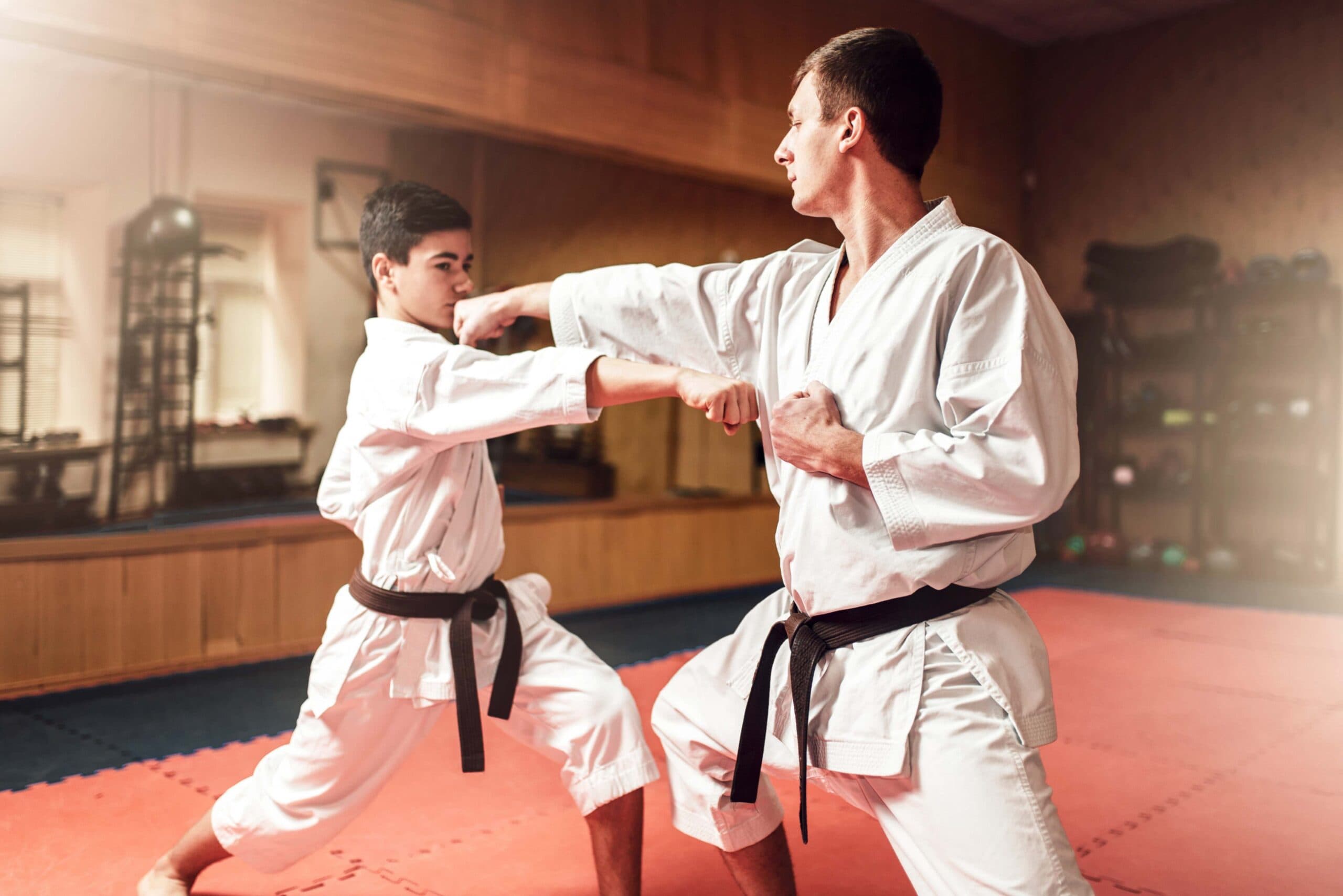 Alt text: Karate instructor demonstrating a straight punch technique with a young student during a training session in a martial arts dojo, both wearing white gis and black belts.