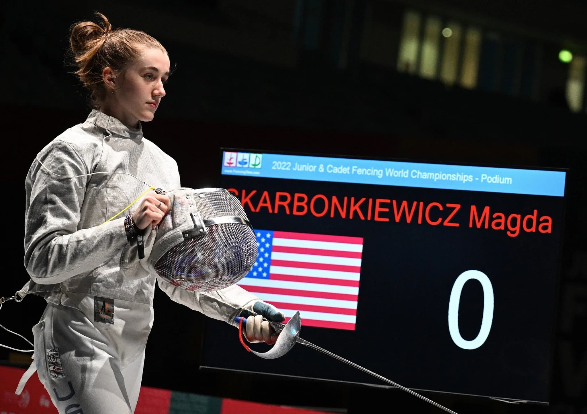 Fencer in full protective gear holds a mask and épée while standing near a scoreboard displaying her name and a U.S. flag during a competition.