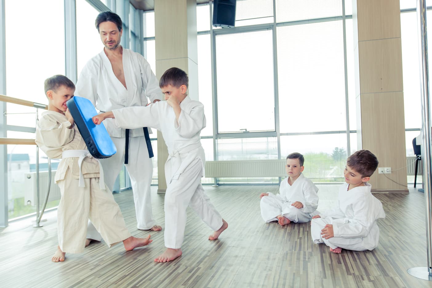 Martial arts instructor supervising children practicing punching techniques with a training pad in a bright studio.