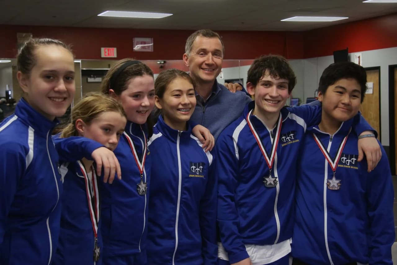 Group of smiling youth athletes wearing matching jackets and medals stand with their coach in a gym, representing a successful team supported by gym membership software.